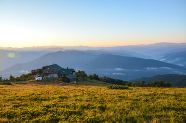 Fototapeta premium Serene countryside landscape featuring house amidst rolling hills during sunrise. View shows expansive greenery and gentle mountains Peaceful and scenic this setting invites tranquility and relaxation