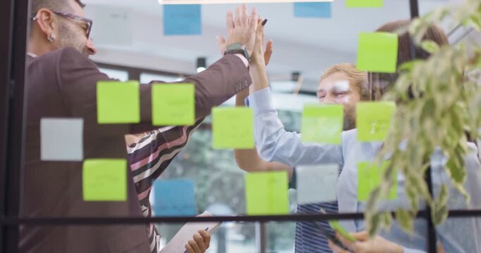 Work colleagues discussing startup project ideas and giving high five. Team of caucasian business people at glass wall with sticky notes. Scrum with colorful stickers. Low angle view.