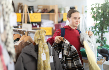 Teenage girl is choosing winter outerwear with interest at a sale in a store, she is holding hangers with jackets and coats