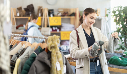 Adult woman stands in a clothing store against the background of hangers and chooses knitted hats for winter. Visitor to the boutique makes purchases at discounts