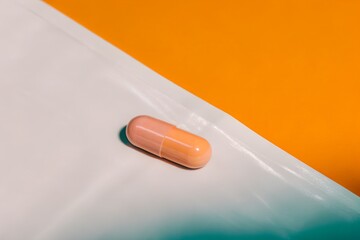 Single Peach Colored Capsule on a White Surface with Bright Orange Background and Harsh Shadow. Peach capsule on a white surface and orange background