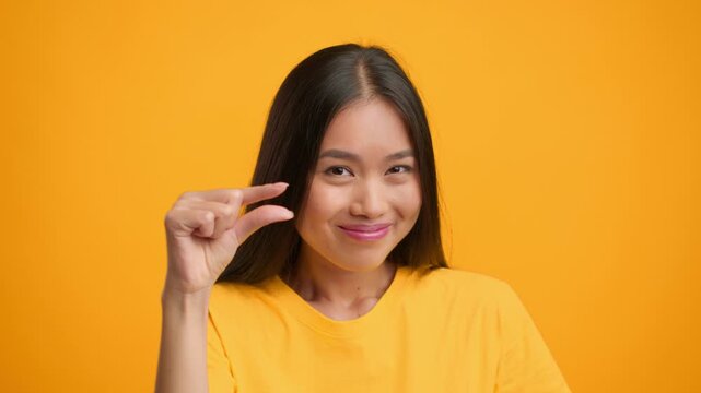 A millennial woman with long hair playfully makes a gesture with her hand, showing a small size. She stands in front of a vibrant orange backdrop, adding energy to the moment.