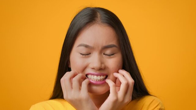 A young Asian woman with long hair displays a frustrated expression, clutching her face with her hands. The bright yellow backdrop emphasizes her emotion, capturing a relatable moment in lifestyle.