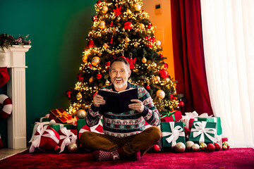 Cheerful elderly man reading a book by the Christmas tree surrounded by gifts, enjoying the festive spirit at home