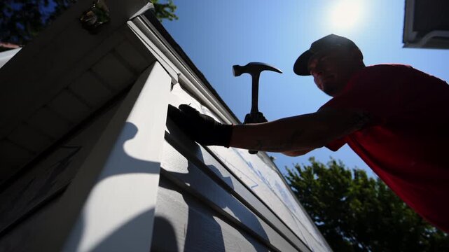 Worker hummers a nail into plastic panel siding on the outside wall of the residential house against bright blue sky