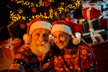 Couple in Santa hats smile beside a decorated Christmas tree with gifts in a warm indoor festive home setting tonight