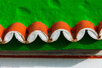 Orange barrel roof tiles with white undersides contrasting against green stucco wall. Curved red and white roof tiles form wave pattern against wall. Scalloped relief of curved clay roofing elements