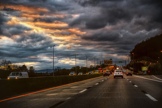 A dramatic sky with dark, fiery clouds hangs over a busy highway during the evening commute. The image captures the movement of vehicles on the interstate with bright orange and white lights.