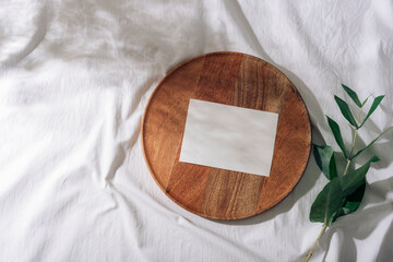 Wooden plate with blank card on white fabric with green leaves in sunlight. Holiday greeting card. Top view, flat lay, mockup