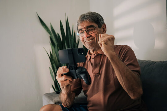 Elderly man celebrating win while playing mobile game on smartphone controller at home - Powered by Adobe
