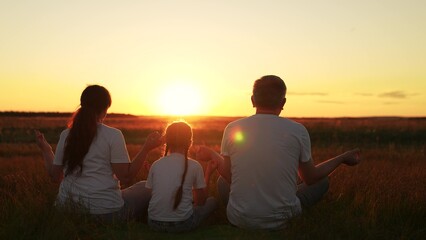 Happy family sits on lawn doing yoga in lotus position raising their arms up in sunset park. Father child mother practice relaxing yoga outdoors lawn. People meditate in nature. Sports, people, health