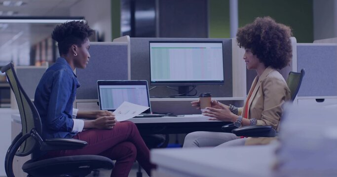 Holding reports and coffee cup, two colleagues reviewing spreadsheet data in open-plan office