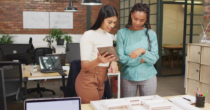 Colleagues wearing office attire using tablet while reviewing scale model at desk, with laptops