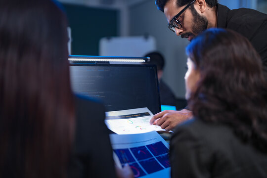 A senior data scientist with glasses points out an anomaly in a machine learning model's code to two female AI engineers, as they finalize the algorithm for a new predictive analytics service.
