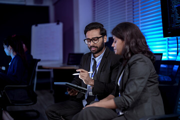 A diverse team of professionals collaborates on a business strategy late at night. A male manager uses a tablet and stylus to explain key points to his female colleague in a modern boardroom.