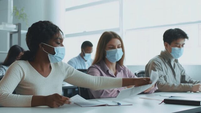 In a bright classroom, diverse college students wear masks while collaborating on a project. They exchange notes and ideas, emphasizing teamwork and education in a university setting. - Powered by Adobe