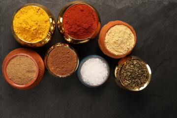 Overhead top view flat lay colourful array of spices herbs powder blue clay ceramic brass metal pot on dark black slate surface with a clean and neat appearance spice copy text space border frame