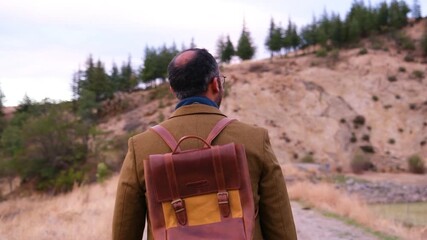 Rear view of middle-aged man in brown jacket and leather backpack walking slowly along rural path in slow motion - Powered by Adobe