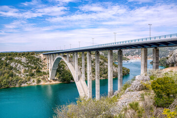 View of the Krka river in Croatia with a large concrete bridge, turquoise water, rocks and green vegetation under blue sky.