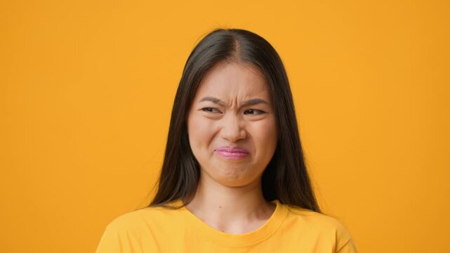 A young Asian woman displays a strong expression of distaste or confusion against a vivid yellow background. She is styled casually, reflecting a modern lifestyle.