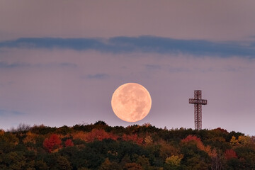 A supermoon rises above autumn treetops and a cross, with a radio tower silhouette. g.