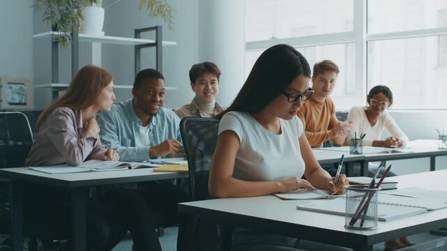 Diverse group of students bullying their classmate Asian lady wearing eyeglasses sitting in front, classroom interior