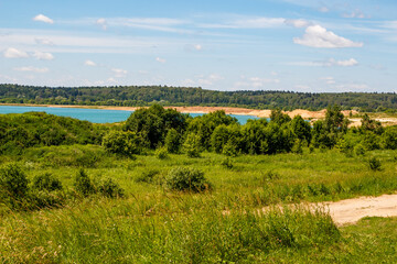 Obraz premium Vibrant green summer landscape with a tranquil azure lake. Lush foliage and a winding dirt path under a clear blue sky