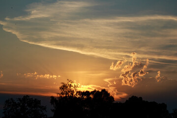 Fiery sunset paints the sky with warm hues, casting silhouetted trees against a glowing horizon. Dramatic clouds catch the golden light, creating a breathtaking, tranquil spectacle
