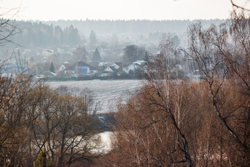 Bare birches and reddish foliage frame a distant village in a hazy, muted landscape. A river winds through the partially snow-dusted field below