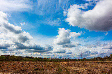 Fototapeta premium Wide open blue heavens, adorned with voluminous, dynamic clouds. A parched field with a track extends towards distant greenery and dwellings