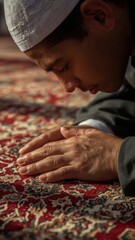 Fototapeta premium Young boy bowing his head in prayer on a richly patterned carpet wearing a white kufi