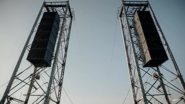 Two massive speaker arrays on towering metal structures against a bright sky, cityscape in background