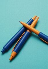 Two Blue and Orange Pens Crossed on Teal Textured Surface Studio Shot