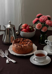 Homemade tea party - apple sponge cake, coffee pot, cups, flowers asters on a round table with a brown tablecloth