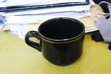 A black ceramic cup placed on a wooden desk with stacks of papers in the background.