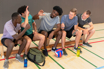 Diverse male volleyball players chatting and high-fiving on bench at gym with ball, duffel bag