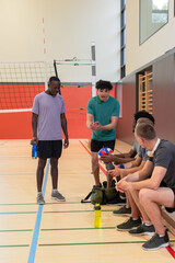 Diverse male teammates gathering beside volleyball net in gym holding water bottles, volleyballs