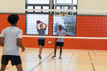 Diverse male partners serving volleyball across net on volleyball court with basketball hoop
