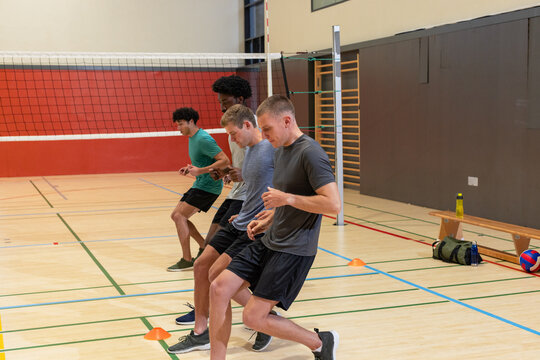 Diverse male players practicing footwork drills at gym using orange cones beside volleyball net - Powered by Adobe