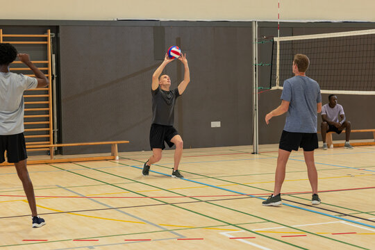 Diverse male volleyball partners in sportswear warming up around net in school gym with volleyball