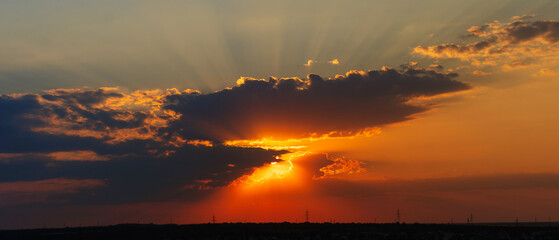 A radiant sunset shows golden sun rays breaking through the edges of dark clouds, illuminating the orange sky above a silhouetted horizon with power lines