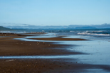 A wide sandy shoreline with gentle waves reflects the sky, while seabirds gather along the coast...