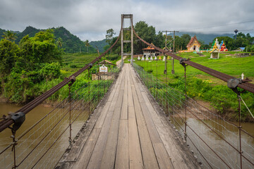 A suspension bridge crossing the Khwae Noi River to Wat Hat Ngio Rai. It offers scenic views of the river, mountains, and temple in Sai Yok District, Kanchanaburi, Thailand.