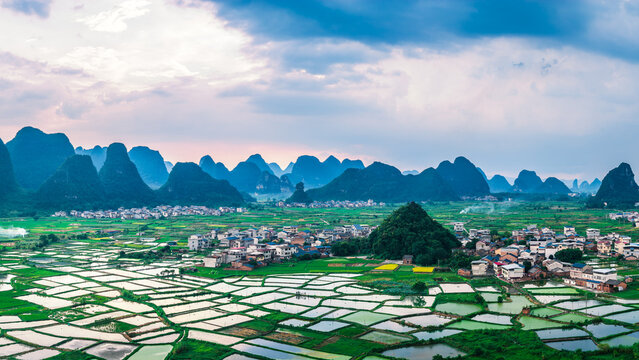 Scenic view of a traditional village surrounded by reflective rice paddy fields at the base of karst mountains at sunset.