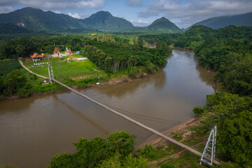 A stunning drone view of the suspension bridge over the Khwae Noi River leading to Wat Hat Ngio Rai, offering panoramic sights of the river, mountains, and temple in Sai Yok, Kanchanaburi.