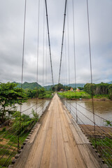 Obraz premium A suspension bridge crossing the Khwae Noi River to Wat Hat Ngio Rai. It offers scenic views of the river, mountains, and temple in Sai Yok District, Kanchanaburi, Thailand.