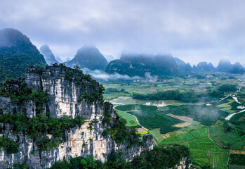 High cliff overlooking a vast green valley with farmland and karst mountains shrouded in morning...
