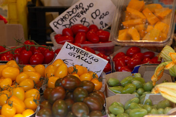 A vibrant display of assorted heirloom tomatoes, including red, yellow datterino, and green varieties, sold in plastic containers at a fresh produce stall in the Rialto Market in Venice.

