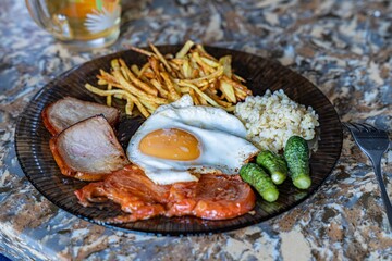 A hearty breakfast plate featuring fried eggs, ham, rice, crispy fries, and pickles, served on a decorative black plate.