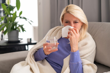 Middle aged blonde woman sitting on couch, blowing her nose due to cold.